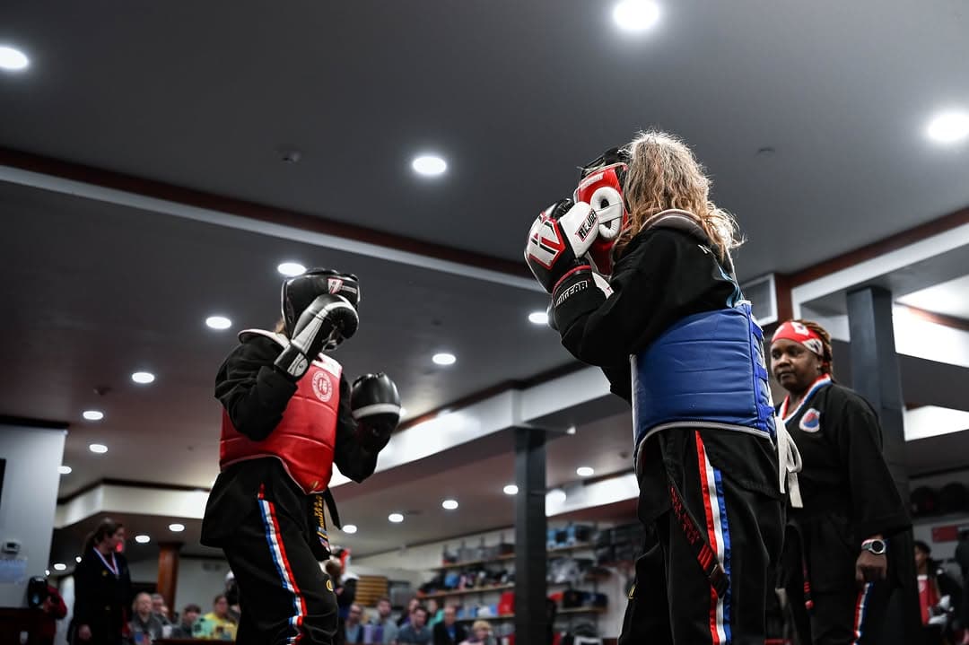 Two students sparring in headgear and chest guards while spectators watch