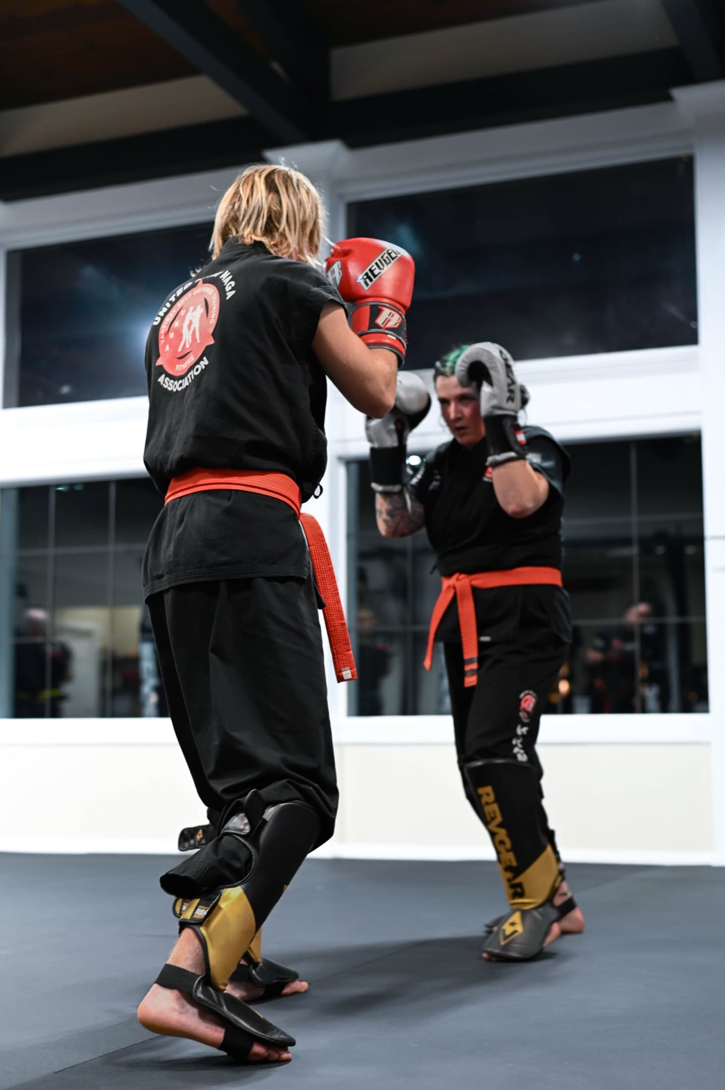 Two adults sparring in black uniforms with gloves and shin guards