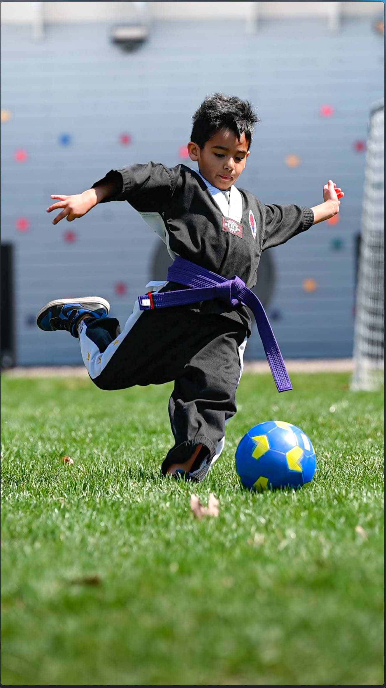 Boy in a dobok kicking a football outdoors with the climbing wall behind